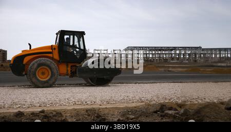 Rouleau jaune compresse le gravier sur un chantier de construction avec une structure de bâtiment industriel à ossature d'acier en arrière-plan. Banque D'Images