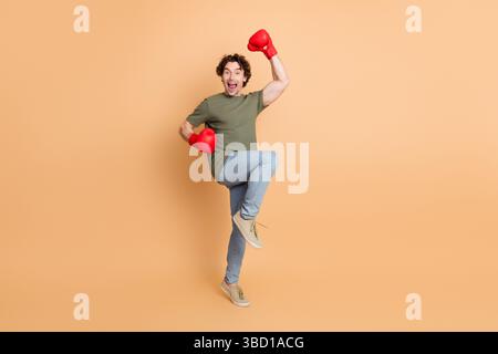 Un jeune homme excité portant un style décontracté et des gants de boxe rouges célèbre la victoire avec un geste joyeux sur fond beige Banque D'Images