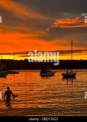 Coucher de soleil orange saisissant sur les eaux calmes du port avec des voiliers ancrés et la silhouette d'un homme pataugant, créant une scène côtière tranquille Banque D'Images