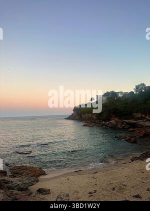 Crique de sable isolée avec de douces vagues, un littoral rocheux et une végétation luxuriante sous un ciel pastel coucher de soleil le long de la côte. Banque D'Images