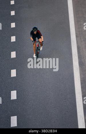 Femme athlétique en équipement de cyclisme aérodynamique roule seule entre les lignes de voie sur l'autoroute de la ville, en maintenant le contrôle et l'équilibre. Banque D'Images