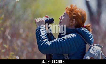 Une femme portant une veste bleue et une queue de cheval rouge regarde à travers une paire de jumelles Banque D'Images
