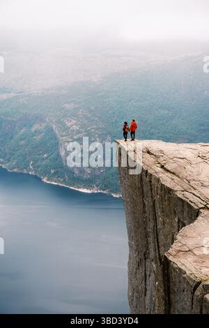Deux aventuriers se tiennent sur le bord de Preikestolen Pulpit Rock, surplombant le fjord ci-dessous. La brume enveloppe les montagnes, créant une atmosphère mystique parfaite pour les amoureux de la nature et les amateurs de sensations fortes. Banque D'Images