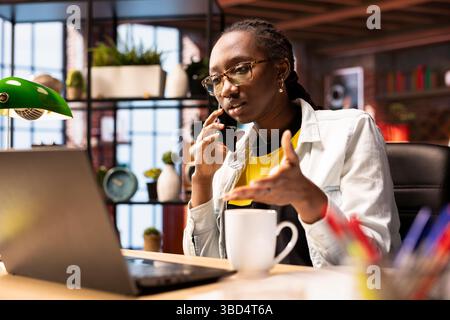 Employé distant dans le bureau à domicile assis au bureau devant l'ordinateur portable, répondant à un appel téléphonique. Femme afro-américaine utilisant un ordinateur portable pour faire du travail en ligne, recevant un appel téléphonique sur smartphone Banque D'Images