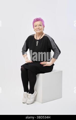 Une vieille femme souriante avec des cheveux roses courts en robe tricotée longue se trouve directement sur une boîte blanche dans le studio Banque D'Images