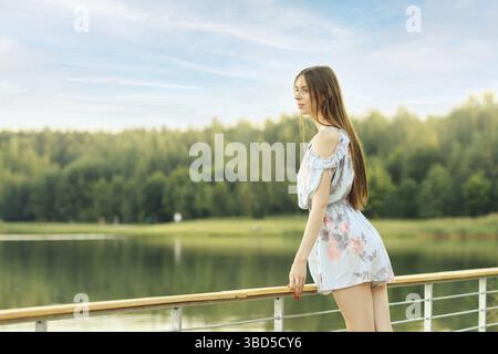 Jeune femme se tient près d'une balustrade, surplombant un lac calme et pittoresque entouré d'une végétation luxuriante, incarnant un moment de paix et de réflexion Banque D'Images