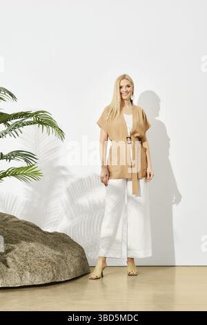 Femme élégante en blouse beige sans manches et pantalon blanc entourée d'éléments naturels comme un rocher et une plante Banque D'Images