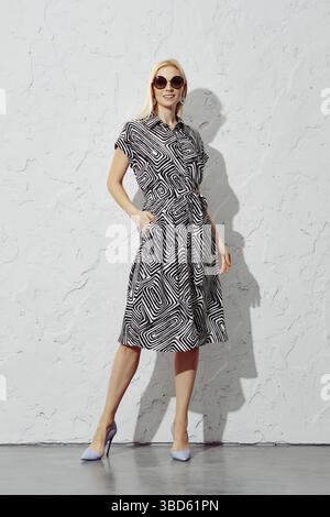 Une femme blonde souriante vêtue d'une robe à motifs et de lunettes de soleil se tient en toute confiance aux rayons du soleil, regardant directement l'appareil photo Banque D'Images