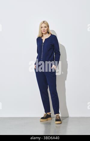 Femme blonde posant dans un pantalon bleu tricoté contre un mur blanc Banque D'Images