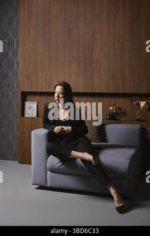 Portrait d'une femme attrayante en costume noir, assise dans un fauteuil dans un appartement moderne Banque D'Images