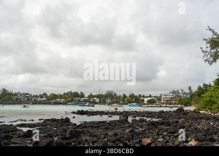 Le Morne, Maurice - 12 mai 2024 : vue panoramique sur le rivage rocheux avec des bateaux ancrés, village et végétation luxuriante sous ciel nuageux Banque D'Images