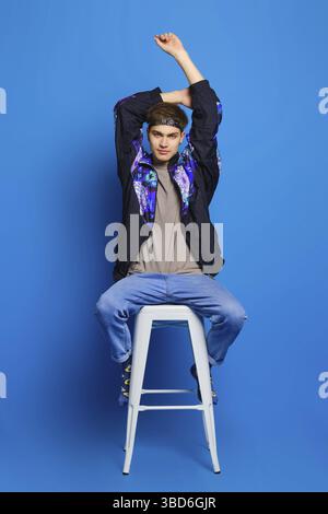 Jeune homme en veste coupe-vent, chemise olive, jeans et chaussettes avec imprimé se dresse sur une chaise haute sur fond bleu studio Banque D'Images