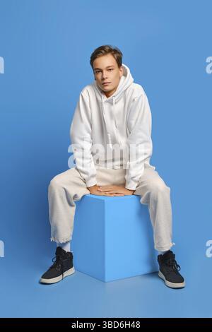 Homme gai avec un sweat-shirt blanc et un pantalon de survêtement assis sur un cube en bois sur fond bleu de studio Banque D'Images