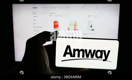 Dans cette illustration photo, une personne tient un téléphone portable avec le logo de la société américaine de marketing multi-niveaux Amway Corp. sur l'écran devant la page Web. Banque D'Images
