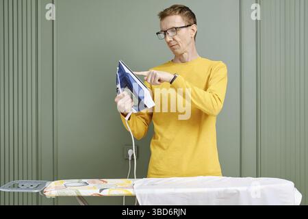 Homme d'âge moyen dans des lunettes touchant la semelle d'un fer avec le doigt Banque D'Images