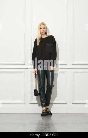 Souriante, une femme élégante pose à l'intérieur portant des bottes en cuir tendance, un pantalon mince et un pull avec ses cheveux blonds à la mode urbaine Banque D'Images