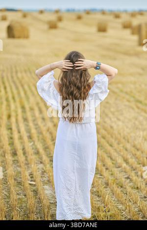 Vue arrière d'une femme en robe blanche et chapeau marchant le long du champ avec des bottes de foin Banque D'Images