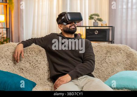 L'homme indien à la maison porte un casque VR sur le canapé et bouge la tête avec étonnement, réagissant au monde virtuel. Hispanic Guy montre de l'excitation et de l'énergie, entièrement immergé dans l'expérience numérique 3D futuriste. Banque D'Images