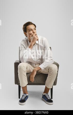 Jeune homme pensif en pantalon beige et en chemise blanche est assis dans un fauteuil en studio Banque D'Images