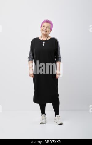 Vieille femme active aux cheveux roses dans une robe en jersey, portrait du corps entier sur un mur blanc Banque D'Images
