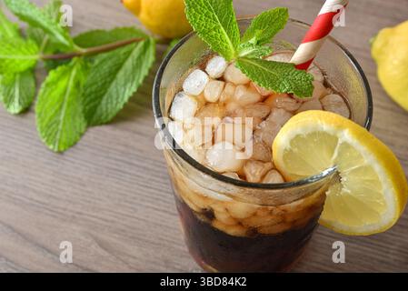 Cocktail à base de cola à la menthe et au citron dans un verre sur une table en bois décorée de glace, feuilles de menthe, citrons et une canette de cola. Vue surélevée de dessus. Banque D'Images