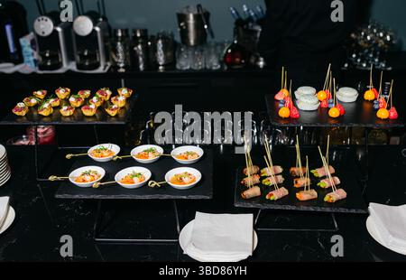 Une table de buffet élégante avec canapés gastronomiques, brochettes de crevettes, bacon roulé, macarons et bouchées de fruits servis sur des plateaux noirs. Banque D'Images