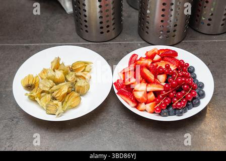 Deux assiettes blanches avec fruits assortis : une avec des fraises tranchées, des bleuets, des groseilles ; l'autre avec des physalis entiers décortiqués. Banque D'Images