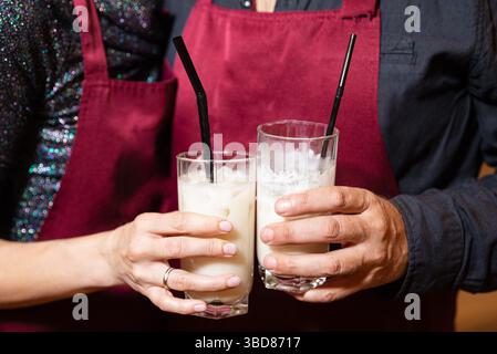 Deux personnes dans des tabliers tenant des verres de cocktails crémeux avec des pailles noires, probablement faites lors d'un cours ou d'un événement de mixologie. Banque D'Images