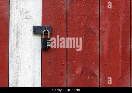 Une porte en bois vieilli peint en rouge et blanc se tient sécurisée avec un cadenas robuste, placé dans un environnement extérieur calme. Banque D'Images