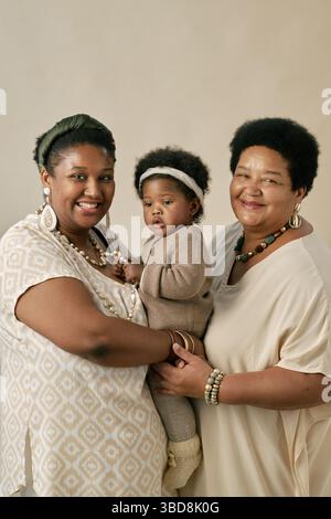 Deux femmes noires avec enfant nourrisson, toutes souriantes et posant pour un portrait de famille tenant l'enfant portant une tenue tricotée, femme à gauche ornée de grand e Banque D'Images
