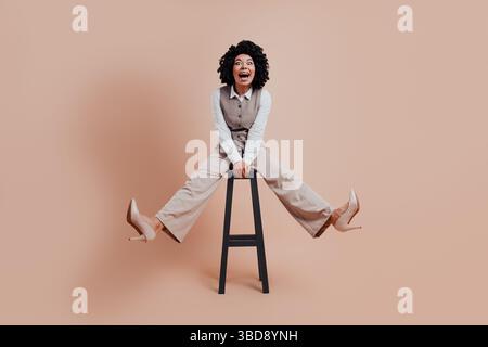 Femme d'affaires confiante dans une tenue élégante se trouve sur un tabouret avec une expression joyeuse sur un fond beige Banque D'Images