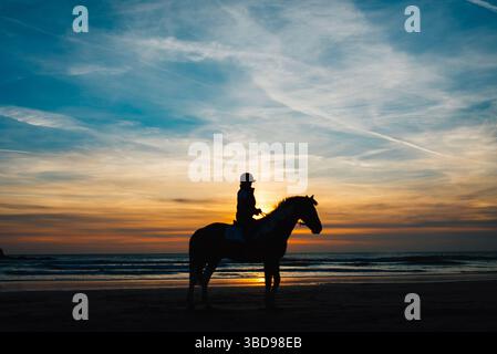 Silhouette d'un cheval et cavalier sur la plage de Perranporth, Cornwall, au coucher du soleil, avec des reflets dorés sur le sable humide et des vagues qui roulent doucement. Banque D'Images