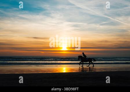 Silhouette d'un cheval et cavalier sur la plage de Perranporth, Cornwall, au coucher du soleil, avec des reflets dorés sur le sable humide et des vagues qui roulent doucement. Banque D'Images