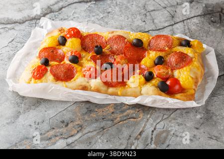 Pinsa romana maison parfumée avec mélange de tomates, saucisse peperoni, olive noire et fromage cheddar gros plan sur papier parchemin sur la table. Horizontal Banque D'Images