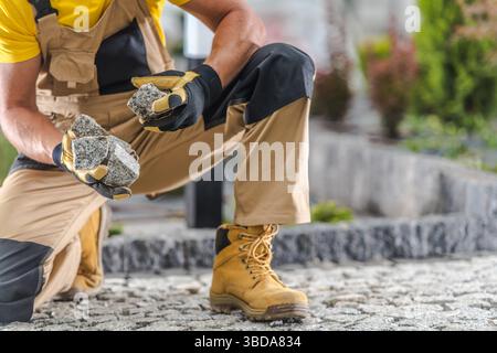 Un ouvrier de la construction s'agenouille sur un chemin de pierre tout en tenant deux pierres dans ses mains. Il porte des gants de protection et des bottes de travail robustes, se concentrant sur Banque D'Images