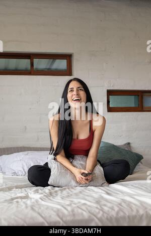 Elle est assise les jambes croisées sur le lit, riant librement, sa joie illuminant la pièce dans un moment de pur bonheur insouciant. Banque D'Images