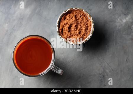 Boisson au chocolat chaud dans une tasse en verre avec poudre de cacao dans un bol bleu sur une surface en béton gris Banque D'Images