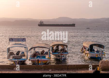 Bateaux touristiques amarrés au coucher du soleil sur la rive d'Aqaba, golfe Persique, avec un cargo et des silhouettes de montagnes israéliennes dans le panorama du moyen-Orient. Banque D'Images