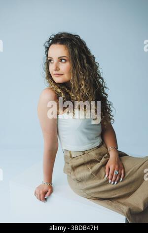 Une jeune femme avec de beaux cheveux bouclés, habillée dans une tenue élégante, s'assoit gracieusement dans un studio minimaliste, exsudant la confiance et mettant en valeur son ch Banque D'Images