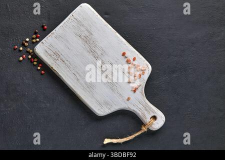 planche à découper en bois blanc et sel rose dispersé et grains de poivre sur une table en béton noir Banque D'Images