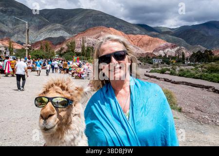 Un touriste pose avec Un lama, les deux portent des lunettes de soleil avec la colline des sept couleurs comme toile de fond, Purmamarca, province de Jujuy, Argentine. Banque D'Images