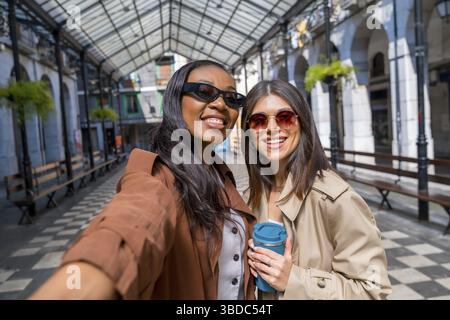 Deux femmes heureuses avec des lunettes de soleil prenant un selfie dans une galerie couverte tout en tenant une tasse de café réutilisable Banque D'Images
