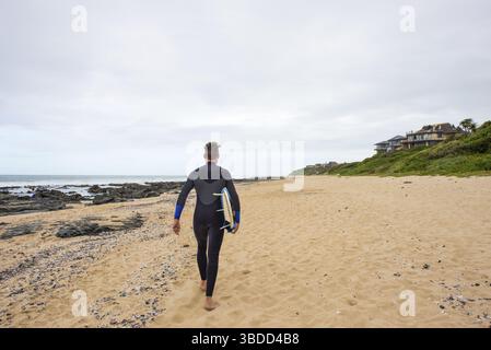 Un surfeur en combinaison marche le long de la plage avec sa planche de surf sous le bras Banque D'Images