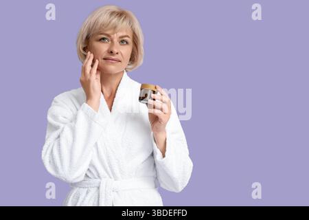 Belle femme heureuse mature en peignoir avec pot de crème sur fond violet Banque D'Images