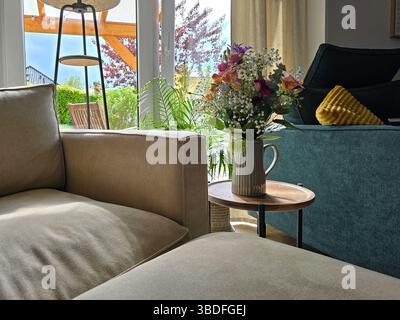 le salon ensoleillé dispose de fauteuils beige et bleu doux, d'une petite table ornée d'un bouquet de fleurs vibrant et offre une vue sur un jardin verdoyant à travers des portes vitrées Banque D'Images