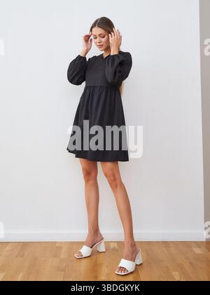 Une femme dans une robe noire à manches longues et des talons blancs se tient sur un plancher en bois. Ses mains sont près de sa tête, et son expression est détendue. La chambre Banque D'Images