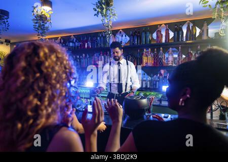 Jeune barman préparant des cocktails pour encourager les clients dans un bar branché avec des lumières bleues et des plantes suspendues Banque D'Images