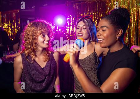 Amies féminines multi ethniques joyeuses chantant karaoké, profitant de la vie nocturne et s'amusant ensemble dans une boîte de nuit Banque D'Images