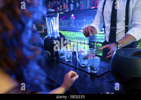 Barman préparant habilement des cocktails bleus éclatants garnis de menthe fraîche et de glace dans une ambiance de boîte de nuit animée Banque D'Images