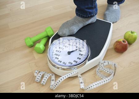 Pieds d'un homme en chaussettes grises marchant sur une balance analogique, haltères verts, ruban à mesurer et pommes couchées à proximité sur un plancher en bois, fitness et Banque D'Images